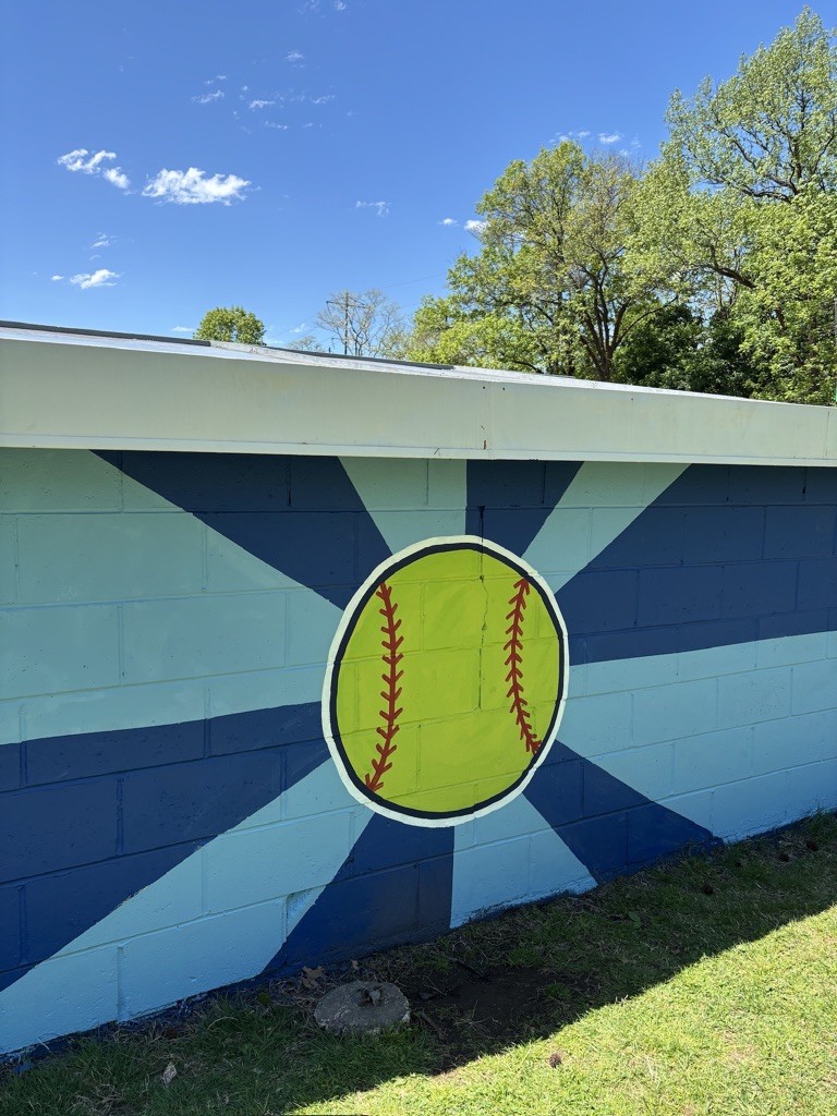 Close-up of a yellow softball with red stitching painted at the center of a blue sunburst wall