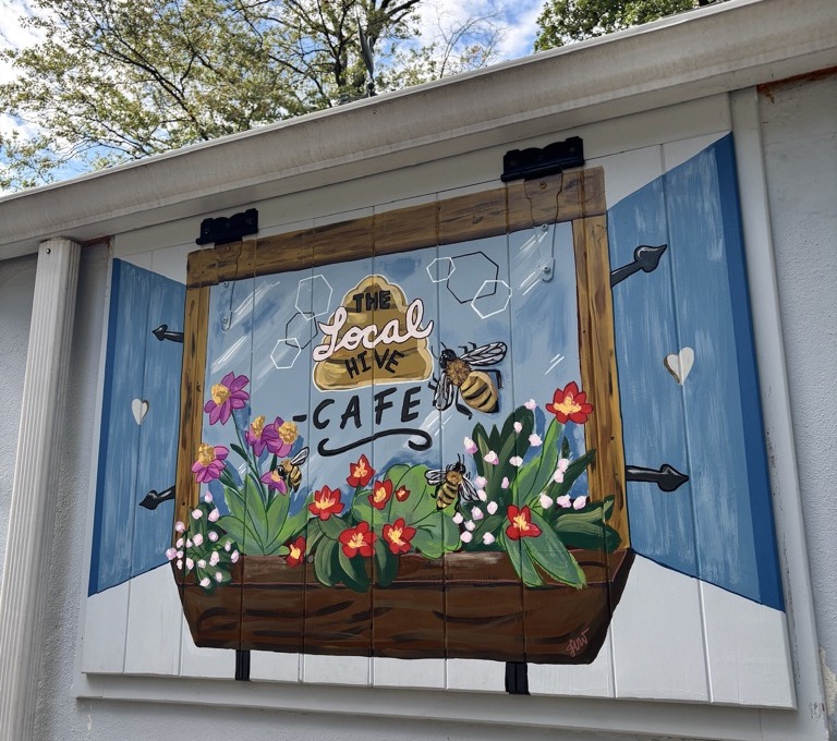 Painted window mural with blue shutters, a flower box of red and pink blooms, buzzing bees and a honeycomb sign reading The Local Hive Cafe