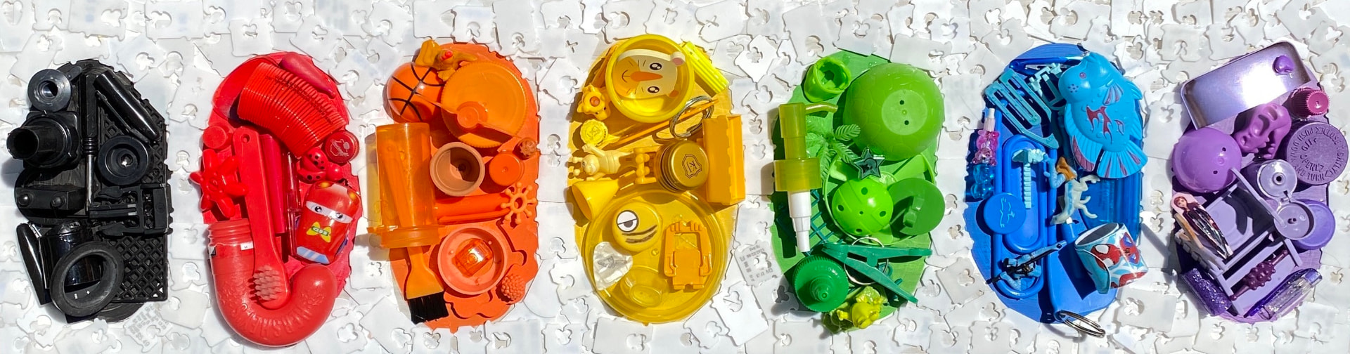 Seven oval clusters of reclaimed plastic toys arranged by color from black through red, orange, yellow, green, blue and purple on white bread tags