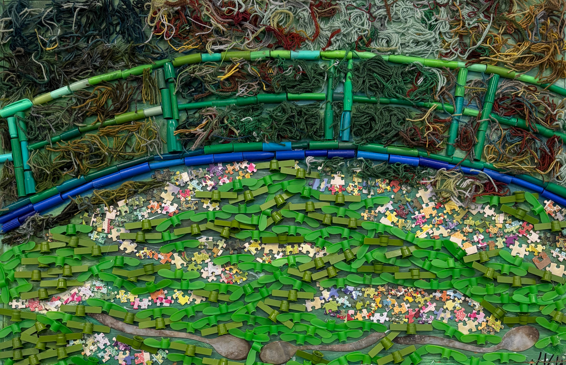 Close-up of a reuse-art panel built from green marker caps, puzzle pieces, yarn, plastic straws and metal spoons forming an Earth Day landscape
