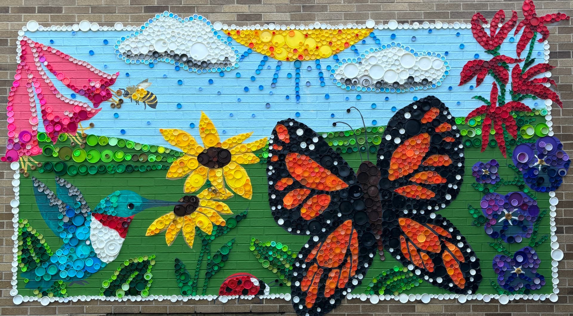 Large outdoor mural of a monarch butterfly, hummingbird, sunflowers and clouds made entirely from recycled plastic bottle caps on a brick wall