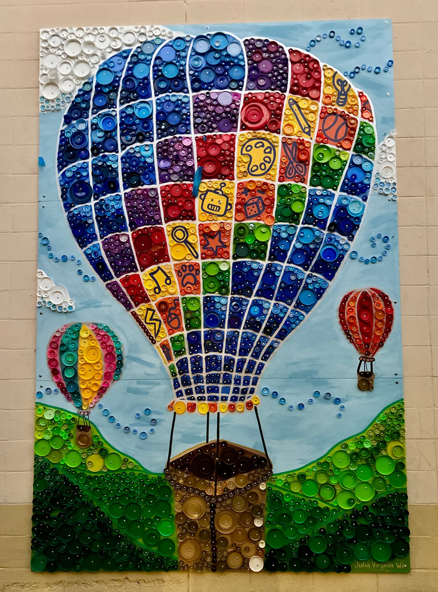 Bottle-cap mural of a giant rainbow hot air balloon decorated with learning icons, floating over green cap hills with two smaller balloons