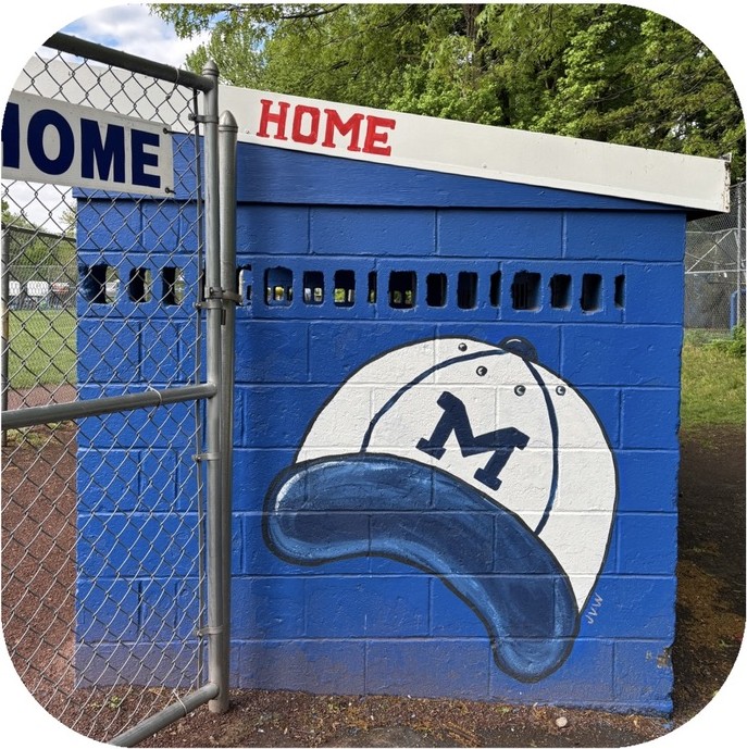 Dugout mural of a white baseball cap with a blue M painted on blue cinder block beside a chain-link gate