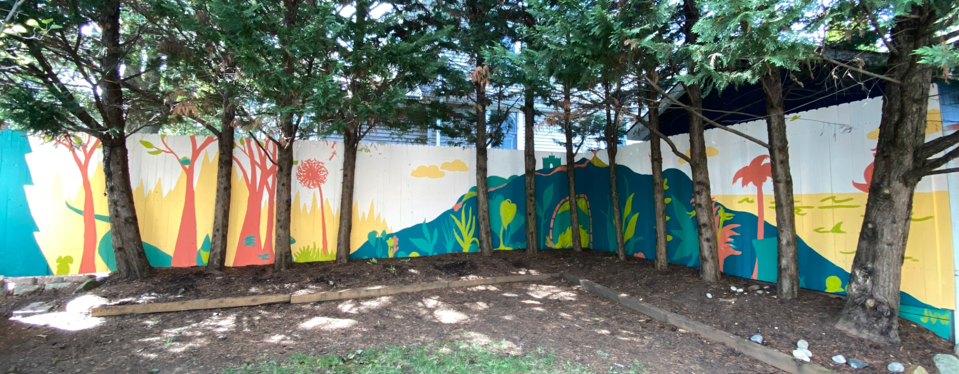 Long wood fence mural of stylized trees, hills, plants and a sailboat in teal, coral and yellow, seen through evergreen trees