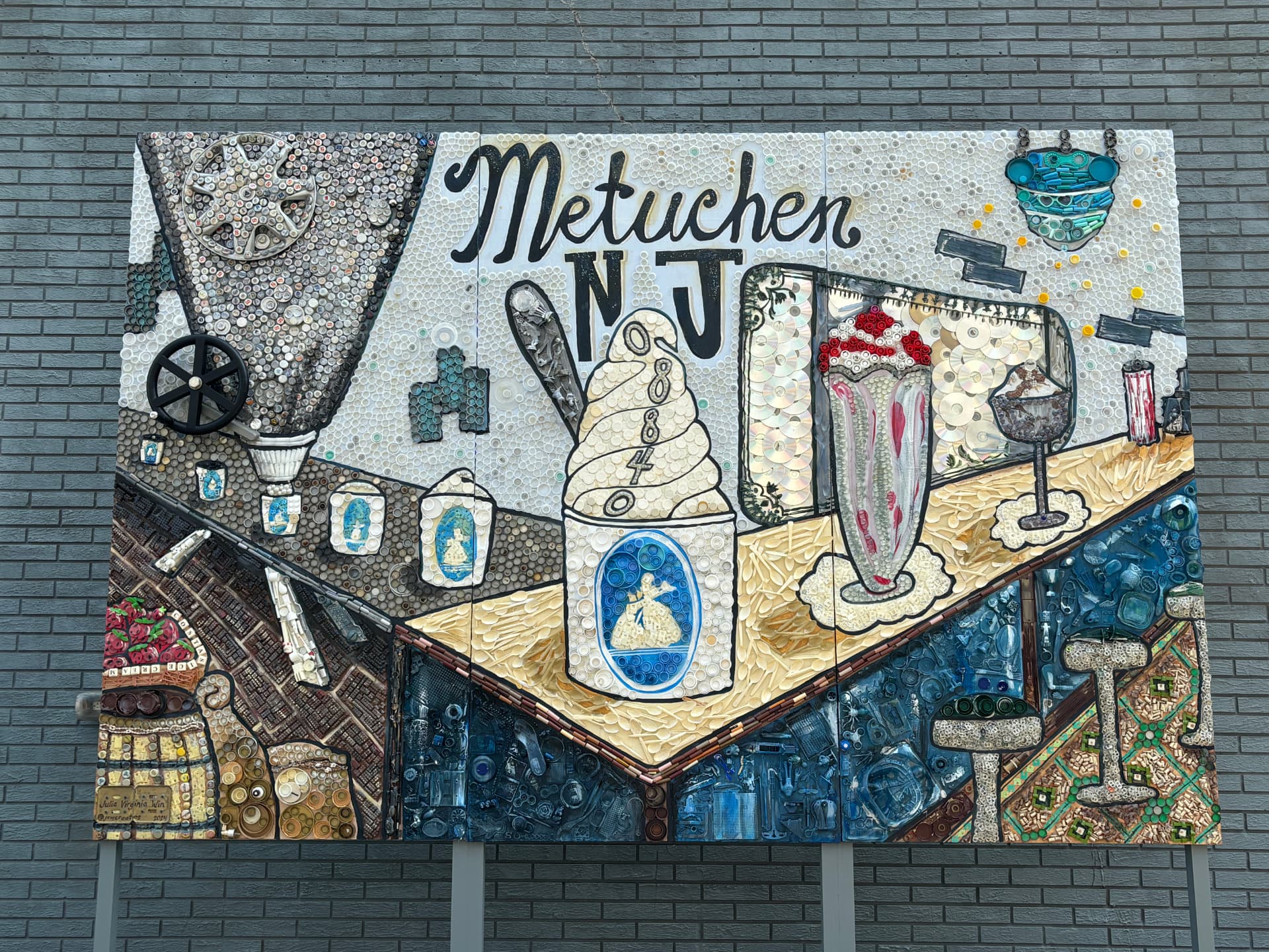 Large wall mural spelling PREserve, built from bottle caps and found objects with an owl, ice cream cone, milkshake, and town icons