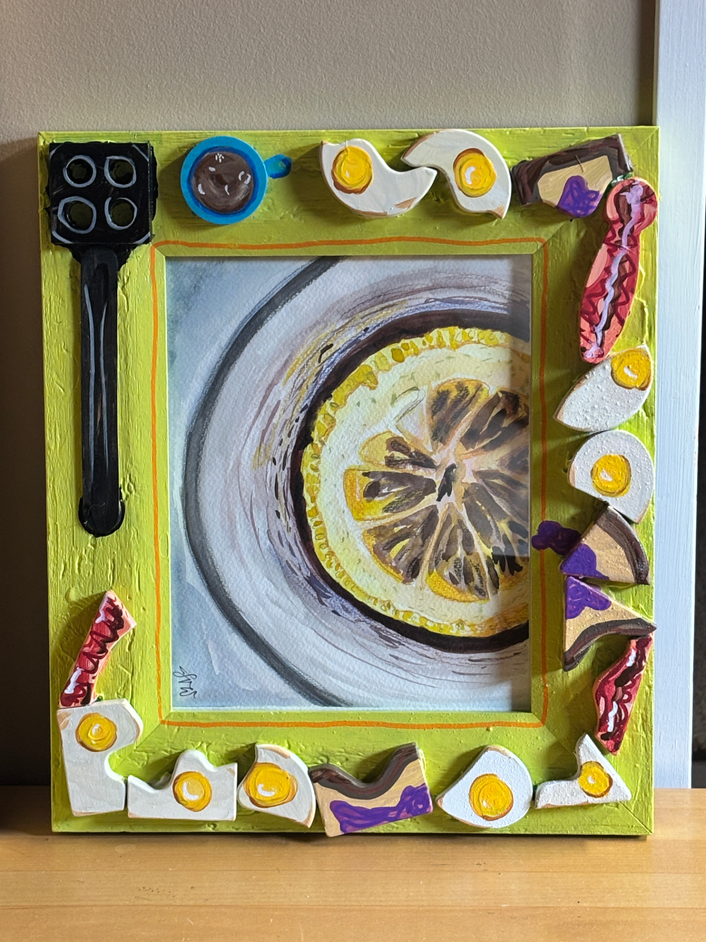Lime-green picture frame trimmed with painted cut-outs of fried eggs, bacon, jam toast, a coffee mug and a black spatula, holding a watercolor of a lemon slice floating in a cup
