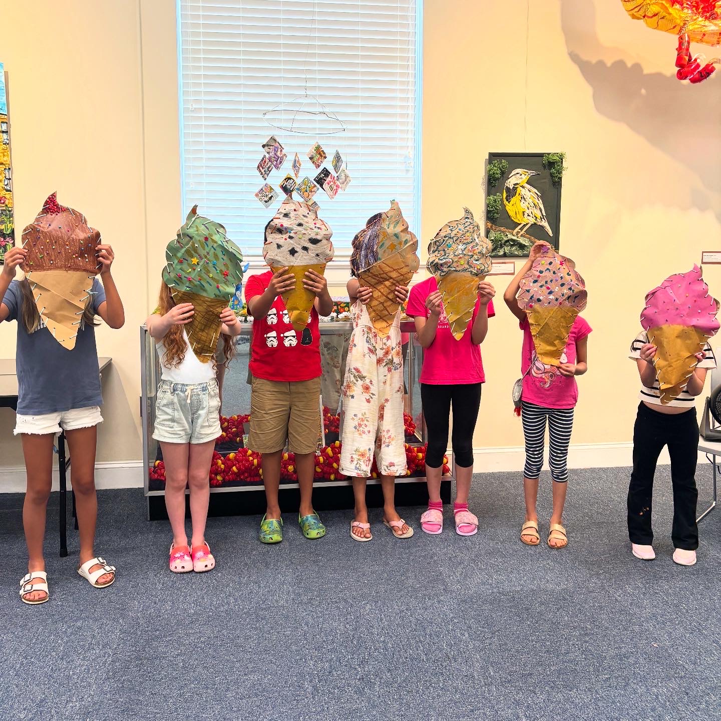 Children at an art workshop holding up giant paper ice cream cone sculptures in front of a bottle cap display