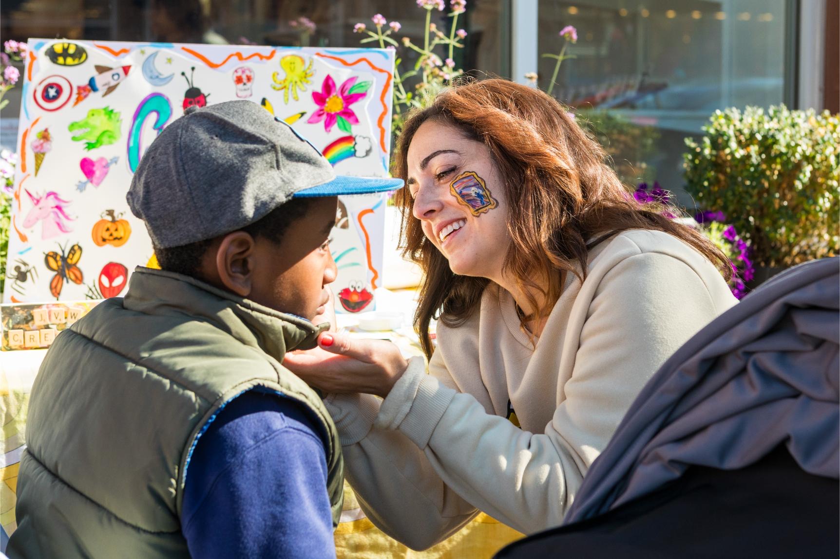Face painter at work on a young boy at an outdoor event, with a board of painted designs on display