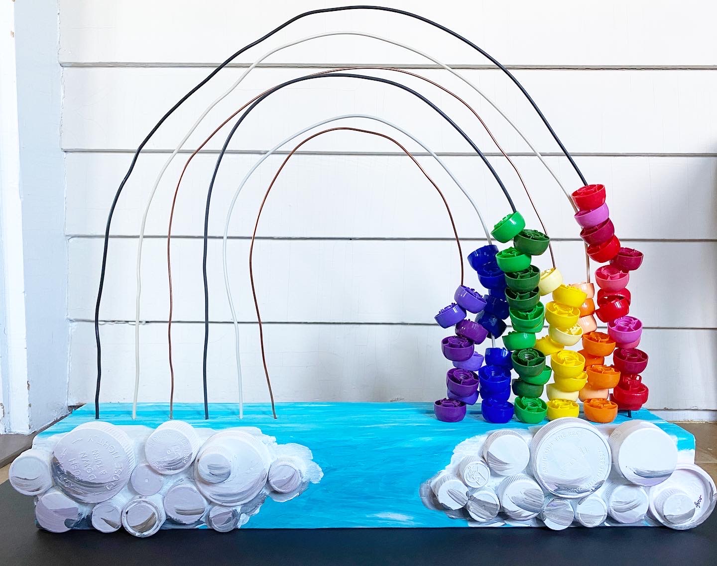 Sculptural rainbow made from stacked colored bottle caps threaded on wire arcs above clouds built from white caps